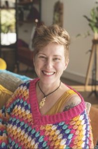 Jacey smiling at you with peachy-orange coloured hair swirled up, wearing a sweater knit in oranges and yellows